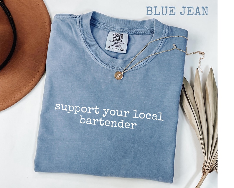 May include: Blue jean colored t-shirt with the text "support your local bartender" in white. The shirt has a crew neck and short sleeves. A gold necklace with a pendant is draped on the shirt.