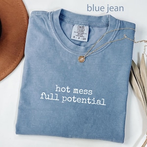 May include: A blue jean colored Comfort Colors t-shirt with the words "hot mess full potential" in white. A gold necklace with a pendant is draped across the shirt. A brown hat and dried plant are in the background.