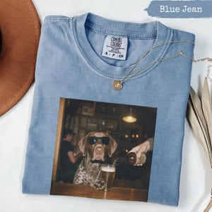 May include: A blue jean colored t-shirt with a graphic print of a dog wearing sunglasses sitting at a bar while a hand pours a drink into a glass. The shirt features a Comfort Colors brand label at the neckline and a small gold pendant necklace.