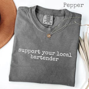 May include: A gray t-shirt with the text "support your local bartender" in white. A gold-colored necklace with a pendant is on the shirt. A brown hat and dried plant are in the background.