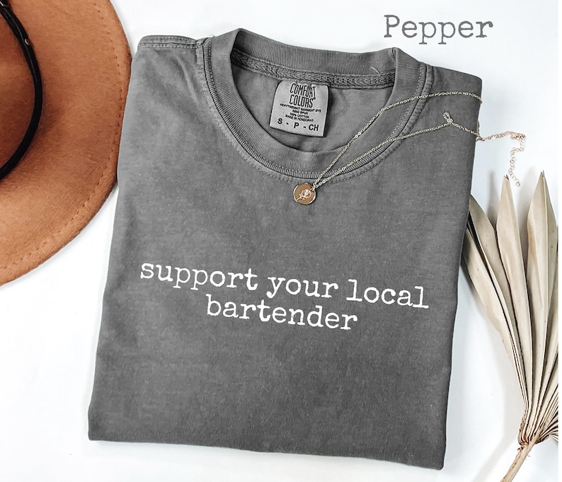 May include: A gray t-shirt with the text "support your local bartender" in white. A gold-colored necklace with a pendant is on the shirt. A brown hat and dried plant are in the background.