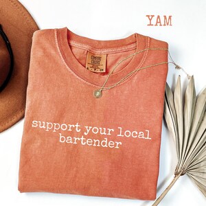 May include: A coral-colored t-shirt with the phrase "support your local bartender" in white lettering. A gold necklace with a pendant is draped on the shirt. A brown hat and a dried palm leaf are in the background.