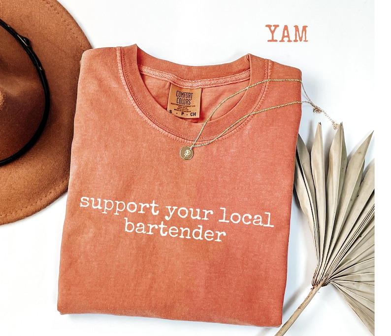 May include: A coral-colored t-shirt with the phrase "support your local bartender" in white lettering. A gold necklace with a pendant is draped on the shirt. A brown hat and a dried palm leaf are in the background.