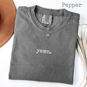 Peut inclure: Un t-shirt gris Comfort Colors avec le mot "yawn." brod&eacute; en blanc. Un collier pendentif dor&eacute; et un chapeau marron sont &agrave; c&ocirc;t&eacute; du t-shirt. Le t-shirt est de taille S-CH.