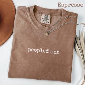 Könnte beinhalten: Espressofarbenes T-Shirt mit dem weißen Schriftzug "peopled out". Das Shirt hat einen Rundhalsausschnitt und ein kleines Etikett am Ausschnitt. Eine silberne Halskette mit einem kleinen Anhänger liegt über dem Shirt. Ein brauner Hut ist im Hintergrund zu sehen.