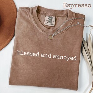 May include: Espresso-colored t-shirt with the words "blessed and annoyed" in white. A silver necklace with a small pendant rests on the shirt. The shirt is a Comfort Colors brand.