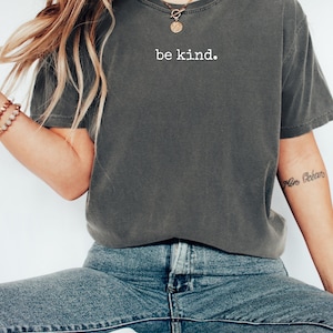 May include: Dark gray t-shirt with the words "be kind" printed in white. The shirt is a relaxed fit and is paired with blue jeans. A gold necklace and bracelets are also visible.