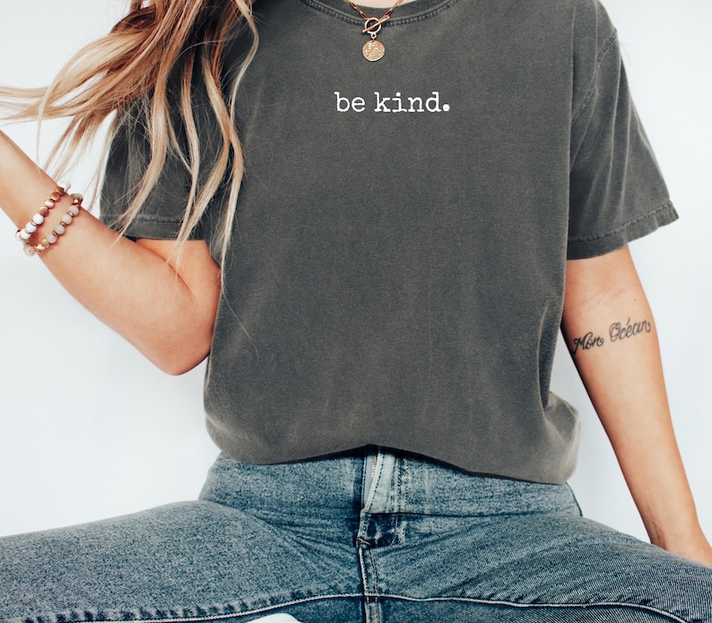 May include: Dark gray t-shirt with the words "be kind" printed in white. The shirt is a relaxed fit and is paired with blue jeans. A gold necklace and bracelets are also visible.