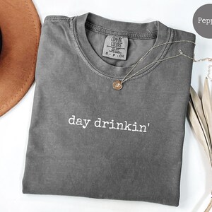 May include: A gray t-shirt with the text "day drinkin'" in white lettering. The shirt is made of a soft, comfortable fabric and has a relaxed fit.