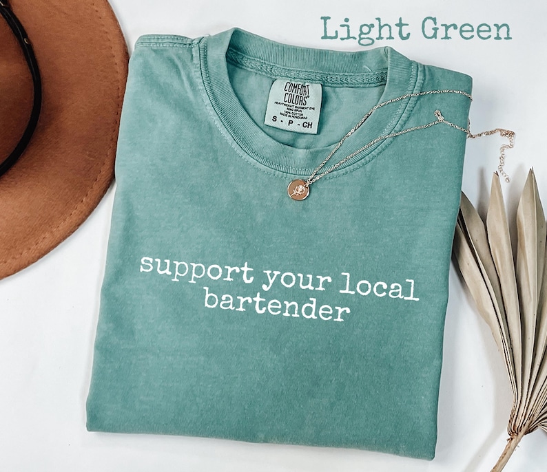 May include: Light green t-shirt with the text "support your local bartender" in white. A gold necklace with a pendant rests on the shirt. The shirt is folded, with a brown hat and dried plant in the background.
