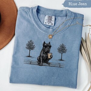 May include: A blue jean coloured t-shirt featuring a graphic of a black dog wearing sunglasses and holding a coffee cup while sitting on a kerb between two trees. The shirt has a Comfort Colors label at the neck and a gold chain necklace rests on top.