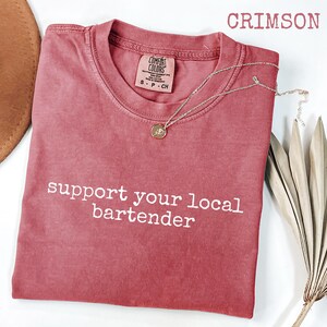 May include: Crimson-colored t-shirt with the text "support your local bartender" in white. The shirt has a crew neck and a small tag on the upper right. A gold necklace with a pendant is draped on the shirt. A brown hat is visible in the upper left.