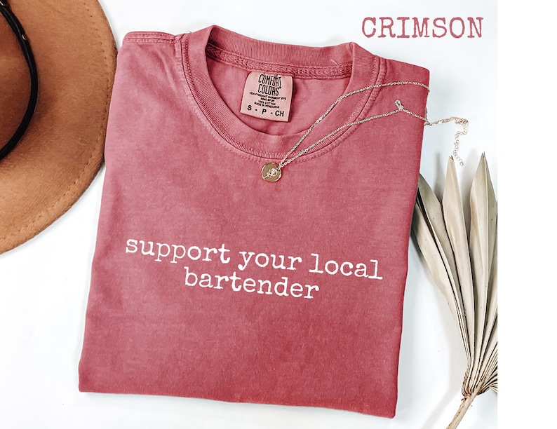 May include: Crimson-colored t-shirt with the text "support your local bartender" in white. The shirt has a crew neck and a small tag on the upper right. A gold necklace with a pendant is draped on the shirt. A brown hat is visible in the upper left.