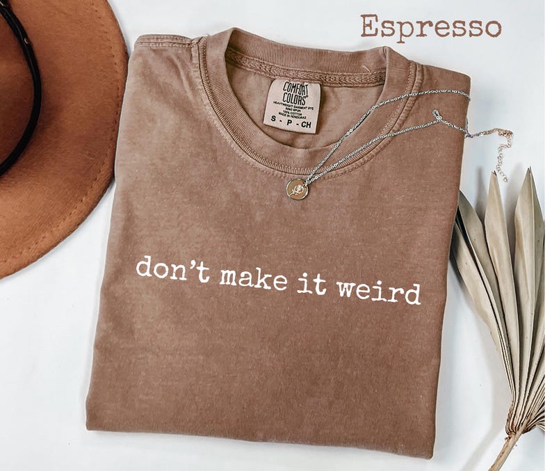 May include: Espresso-colored t-shirt with the phrase "don't make it weird" in white. A silver necklace with a small pendant rests on the shirt. The shirt is a Comfort Colors brand.