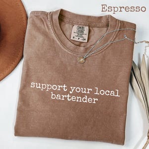 May include: Espresso-colored t-shirt with the text "support your local bartender" in white. The shirt has a crew neck and a small tag. A silver necklace with a pendant is draped across the shirt.