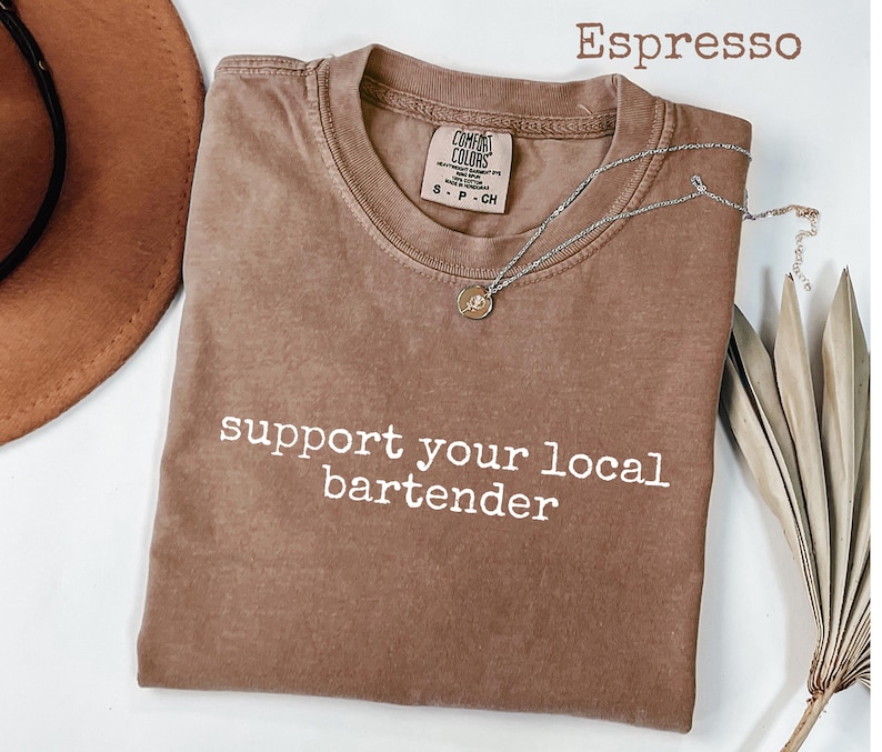 May include: Espresso-colored t-shirt with the text "support your local bartender" in white. The shirt has a crew neck and a small tag. A silver necklace with a pendant is draped across the shirt.