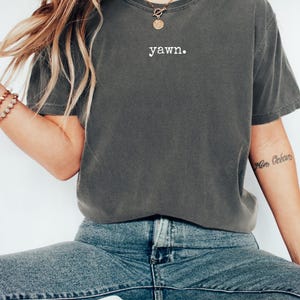 Peut inclure: T-shirt gris fonc&eacute; avec le mot "yawn" imprim&eacute; en blanc. Le t-shirt a une coupe d&eacute;contract&eacute;e et est port&eacute; avec un jean bleu. L'image montre un style d&eacute;contract&eacute; et quotidien.