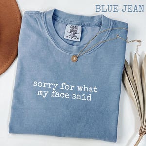 May include: A blue jean colored t-shirt with the text "sorry for what my face said" in white. The shirt has a crew neck and short sleeves. A gold necklace with a pendant is draped on the shirt.