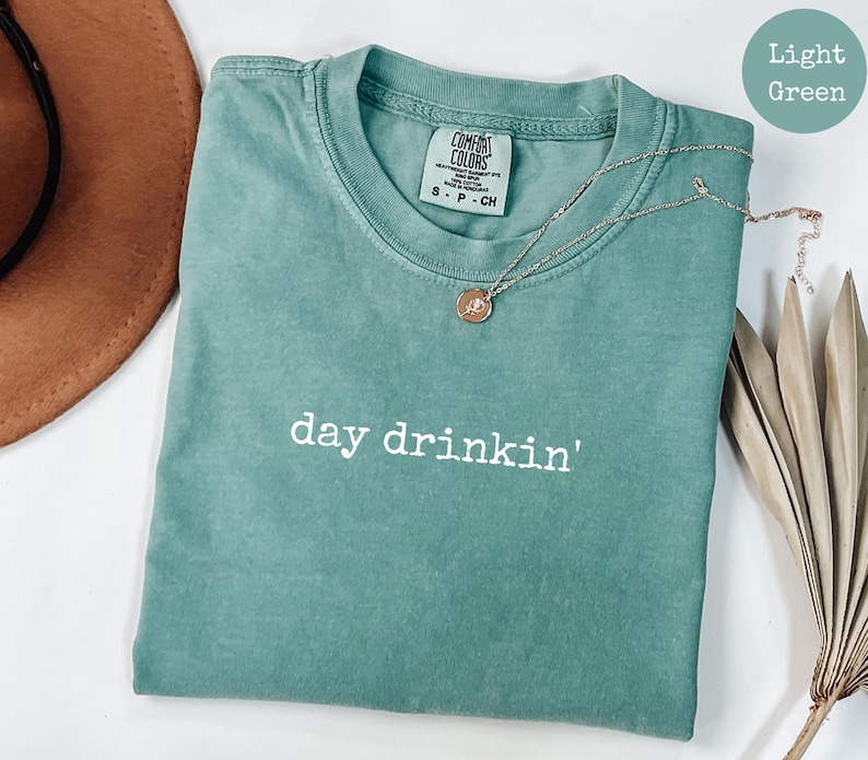 May include: Light green t-shirt with the text "day drinkin'" in white. The shirt is made of a soft, comfortable fabric.