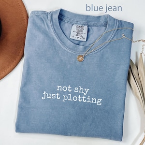 May include: A blue jean colored t-shirt with the text "not shy just plotting" in white. A gold necklace with a small pendant rests on the shirt. The shirt is folded and placed on a white surface.