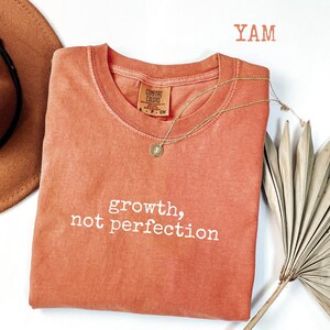 May include: A coral-colored Comfort Colors crewneck t-shirt with the words "growth, not perfection" in white script. A gold necklace with a pendant rests on the shirt. A brown hat and dried palm leaf are in the background.