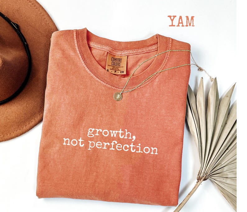 May include: A coral-colored Comfort Colors crewneck t-shirt with the words "growth, not perfection" in white script. A gold necklace with a pendant rests on the shirt. A brown hat and dried palm leaf are in the background.