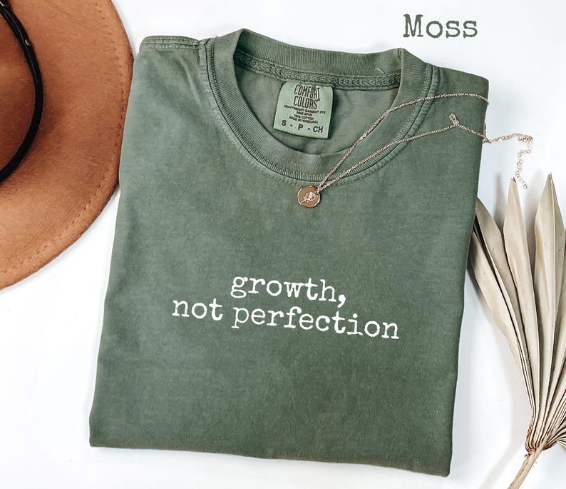 May include: A moss green t-shirt with the words "growth, not perfection" printed in white. A gold necklace with a small pendant rests on the shirt. The shirt is folded, and a brown hat is partially visible.