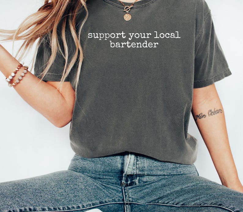 May include: Dark gray t-shirt with the text "support your local bartender" in white. The shirt is a casual style, with a slightly faded appearance. The image shows the shirt on a person wearing blue jeans.