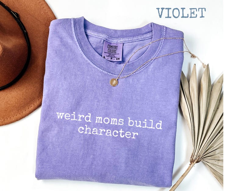May include: A violet Comfort Colors t-shirt with the text "weird moms build character" in white. A gold necklace with a pendant is draped over the shirt. A brown hat is visible in the upper left corner.