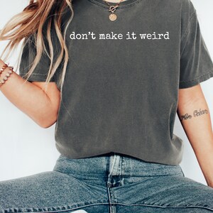 May include: Dark gray t-shirt with the phrase "don't make it weird" printed in white. The shirt is a relaxed fit and has short sleeves. The model is wearing blue jeans and a bracelet.