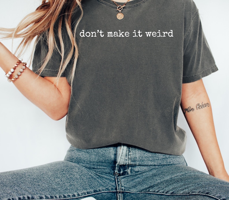May include: Dark gray t-shirt with the phrase "don't make it weird" printed in white. The shirt is a relaxed fit and has short sleeves. The model is wearing blue jeans and a bracelet.