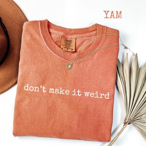May include: A coral-colored t-shirt with the phrase "don't make it weird" in white lettering. A gold necklace with a pendant and a brown hat are next to the shirt. The word "YAM" is in the top right corner.