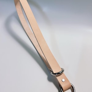 May include: A light tan leather wrist strap with a silver metal ring and a smaller ring attached with a rivet. The strap is made of a single piece of leather and is designed to be worn around the wrist. The text "Chuncui Design Studio" is at the bottom.