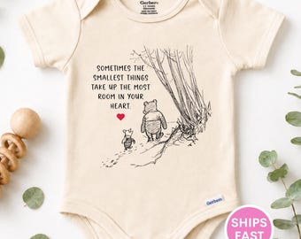 May include: A cream-colored short-sleeve baby bodysuit featuring a black line-art illustration of Winnie the Pooh and Piglet walking near trees. The text reads: Sometimes the smallest things take up the most room in your heart, with a small red heart.