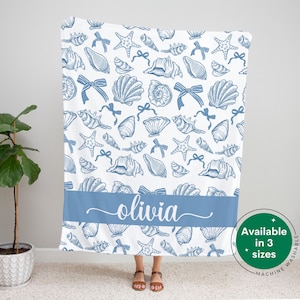 May include: A white fleece blanket with a blue seashell pattern and the name "olivia" printed in blue on a blue stripe across the bottom. The blanket is available in three sizes and is machine washable.