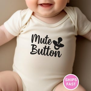 May include: A cream-colored baby onesie with the words "Mute Button" and a pacifier graphic in black. The onesie is short-sleeved and has a round neckline. The baby is smiling.
