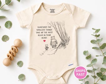 May include: A cream-colored short-sleeve baby bodysuit featuring a black line-art illustration of Winnie the Pooh and Piglet walking. The text reads: SOMETIMES THE SMALLEST THINGS TAKE UP THE MOST ROOM IN YOUR HEART, accompanied by a small red heart.