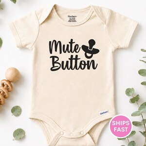 May include: A cream-colored short-sleeve baby bodysuit featuring the black text Mute Button and a graphic of a pacifier. The garment has a round neckline, short sleeves, and snap closures at the bottom. The Gerber brand logo is printed on the inside.