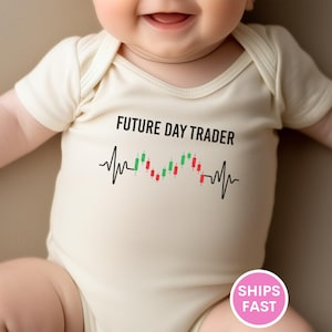 May include: A light beige baby onesie with the text "FUTURE DAY TRADER" in black. Below the text is a graphic of a heartbeat with green and red candlestick patterns. The onesie is short-sleeved.