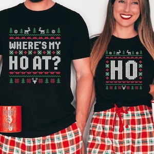 May include: Black t-shirts with a festive design. One shirt reads "WHERE'S MY HO AT?" and the other says "HO". Both shirts have a Christmas sweater-style pattern with reindeer and trees. The models are wearing red and green plaid pajama pants.