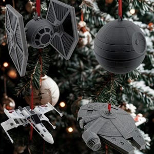 May include: Four grey and white Star Wars-themed Christmas ornaments suspended from red ribbons on a Christmas tree. The ornaments depict a TIE fighter, a Death Star, an X-wing fighter, and the Millennium Falcon.