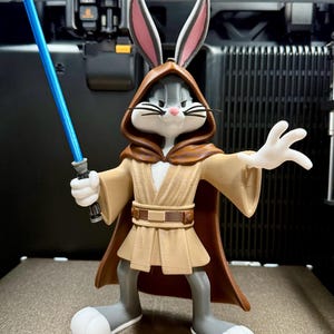 Puede incluir: Figura de Bugs Bunny impresa en 3D vestido de Jedi, sosteniendo un sable de luz azul. La figura lleva una túnica y capucha marrones, con una túnica y cinturón beiges. La figura es gris, blanca y beige, con orejas forradas en rosa.