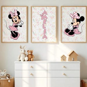 May include: Three framed prints featuring Minnie Mouse in pink dresses and bows, set against a floral background. One print displays the name "Amelia" in pink letters. The prints are displayed above a white dresser with toys.