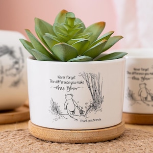Puede incluir: Maceta de cerámica blanca con una ilustración de Winnie the Pooh y el texto "Never forget the difference you make, Miss Glow". Las macetas están sobre un posavasos de madera y una estera tejida. Una maceta contiene una suculenta verde.