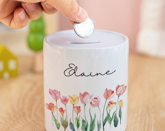 Peut inclure: Une tirelire cylindrique en céramique blanche ornée d'un motif floral de tulipes roses, jaunes et rouges. Le nom "Elaine" est écrit en cursive. Une main insère une pièce. Objet décoratif.