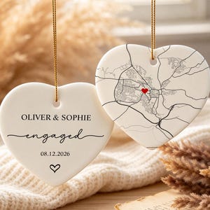 May include: Two white heart-shaped ceramic ornaments hanging from gold strings. One ornament features the text OLIVER & SOPHIE, engaged, and the date 08.12.2026 with a small heart icon. The other displays a black line map with a red heart marker.
