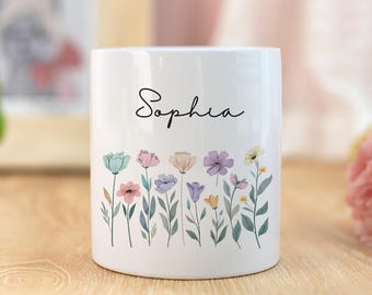 Peut inclure: Mug en céramique blanche avec le nom "Sophia" en écriture cursive. Le mug présente un motif floral coloré au bas, avec des fleurs roses, bleues, violettes et jaunes. Le mug est de forme cylindrique.