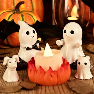 Peut inclure: Deux figurines de fantômes blancs sont assises autour d'une bougie LED centrale en forme de feu de camp avec des flammes orange et rouges. Deux petits fantômes blancs sont placés devant. Les fantômes ont des surfaces lisses et des yeux noirs.