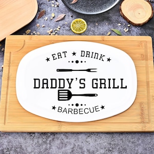 Könnte beinhalten: Weiße ovale Servierplatte mit dem Text "EAT DRINK DADDY'S GRILL BARBECUE" in Schwarz. Die Platte hat ein Gabel- und Spatel-Design. Die Platte liegt auf einem Holzschneidebrett.