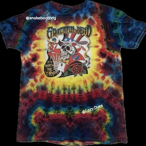 Puede incluir: Camiseta teñida con un diseño de Grateful Dead. El diseño incluye una calavera con sombrero de estrellas y rayas, una rosa y el texto "It's Worth The Trip". La camiseta tiene un patrón de remolino colorido.